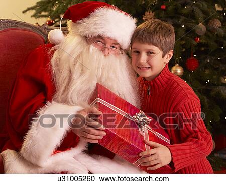 Stock Image - Santa Claus Giving Gift To Boy In Front Of Christmas Tree. Fotosearch