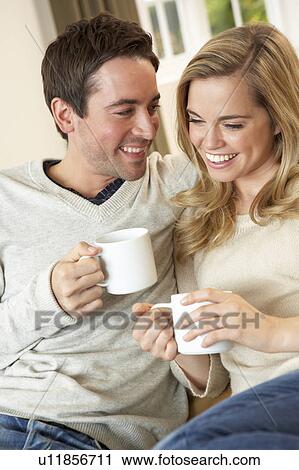 Stock Image - Young couple sitting and relaxing on sofa with cup in hand. Fotosearch