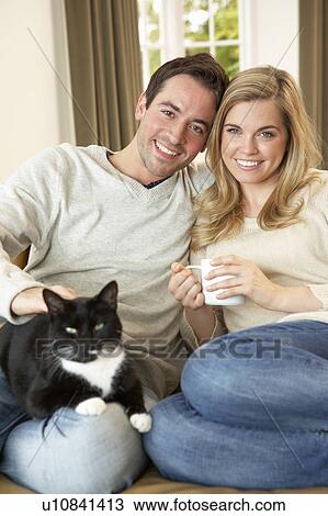 Stock Image - Young couple sitting with cat on sofa with cup in hand. Fotosearch