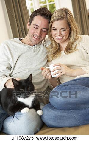 Stock Photography - Young couple sitting with cat on sofa with cup in hand. Fotosearch