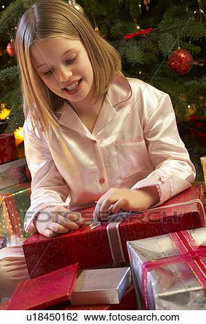 Stock Image - Young Girl Opening Christmas Present In Front Of Tree. Fotosearch