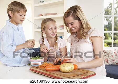 Stock Image - Young mother with children peeling vegetables in kitchen. Fotosearch