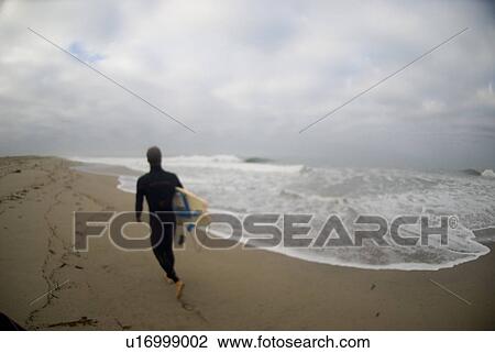 Stock Image - A male walking on beach with surf board. Fotosearch