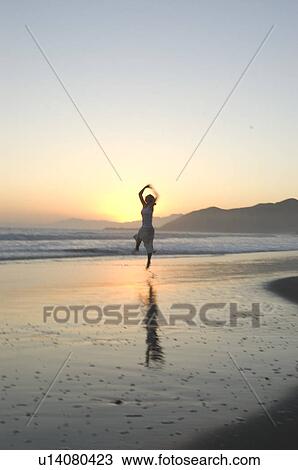 Stock Image - A young female on the beach jumping at sunset. Fotosearch