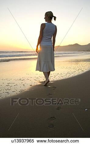 Stock Photography - A young woman walks on the beach at sunset. Fotosearch