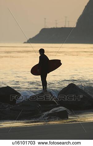 Man holding surfboard standing on rocks at sunset View Large Photo Image Stock Image - Man holding surfboard standing on rocks at sunset. Fotosearch