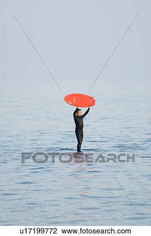 Stock Image - Man standing in the water holding surfboard over his head. Fotosearch