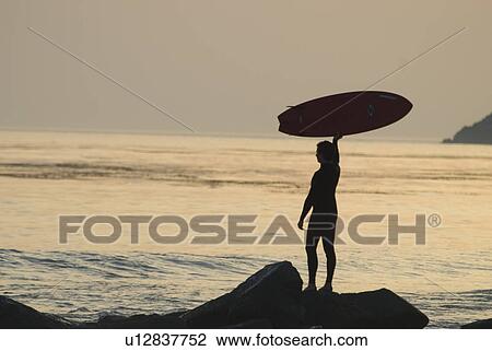 Stock Image - Man with surfboard standing on rocks. Fotosearch
