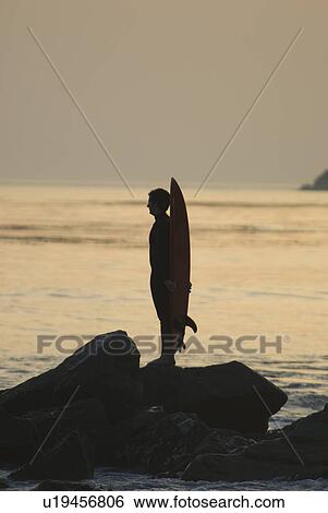 Stock Photograph - Man with surfboard standing on rocks. Fotosearch
