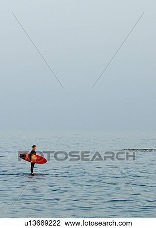 Stock Image - Surfer standing in the water holding surfboard. Fotosearch
