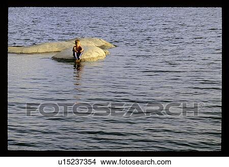 Picture - Boy sitting on a rock in the water. Fotosearch