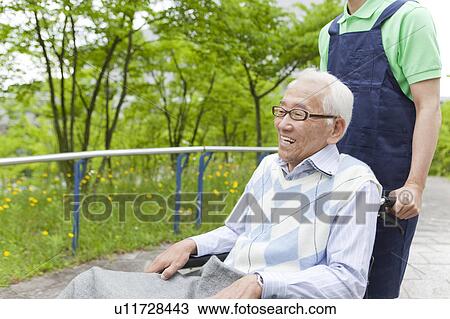 Stock Image - Healthcare worker pushing senior man in wheelchair, Tokyo Prefecture, Honshu, Japan. Fotosearch