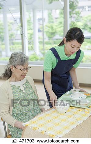 Stock Image - Healthcare worker serving food to senior woman, Kanagawa Prefecture, Honshu, Japan. Fotosearch