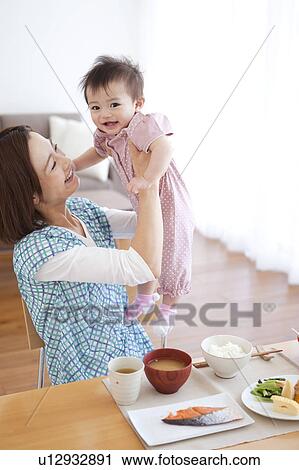Stock Image - Mother Lifting Baby Girl at Breakfast Table. Fotosearch