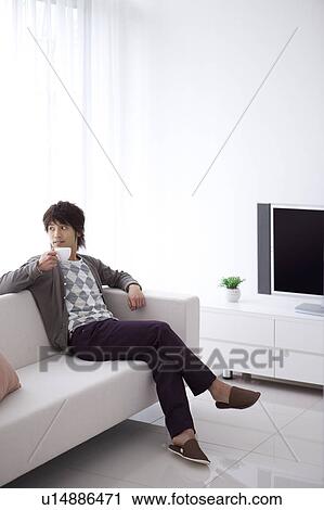 Stock Image - Young man sitting on sofa drinking tea. Fotosearch