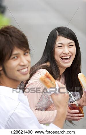 Stock Image - Young couple eating on bench looking at camera. Fotosearch