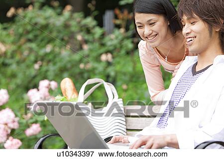 Young couple sitting on a bench using laptop View Large Photo Image Stock Photo - Young couple sitting on a bench using laptop. Fotosearch