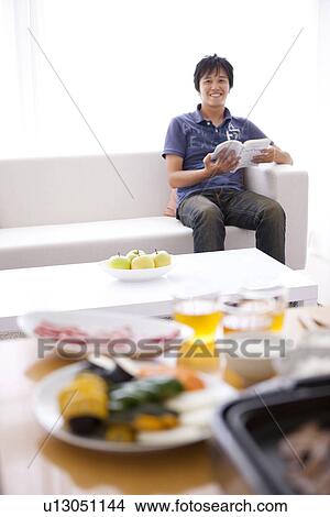 Picture - Young Man Reading Book on Sofa, Dinner on Table. Fotosearch