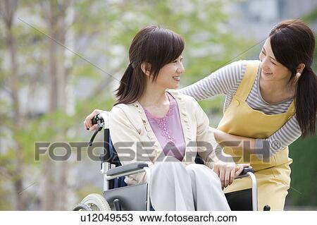 Stock Image - Caretaker and mature woman on wheelchair talking. Fotosearch