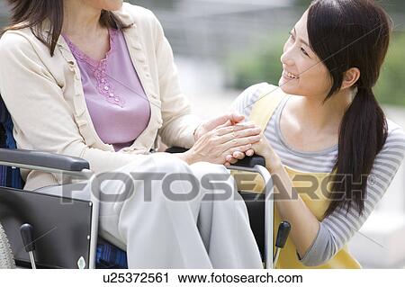 Stock Image - Caretaker and mature woman on wheelchair holding hands. Fotosearch