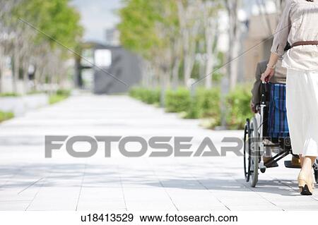Stock Photo - Mature woman pushing senior man on wheelchair, rear view. Fotosearch