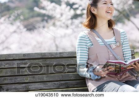 Stock Image - Young woman sitting on bench reading a book. Fotosearch