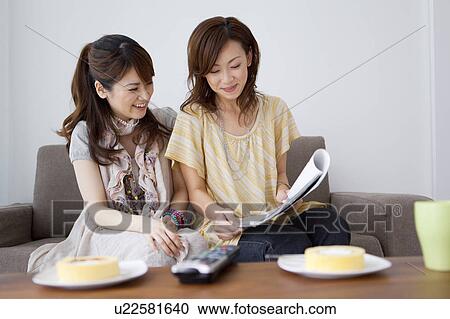 Stock Image - Young woman sitting on sofa reading magazine. Fotosearch