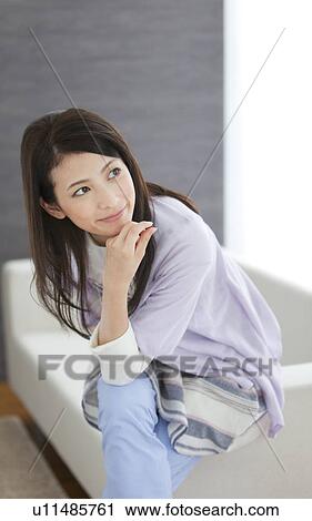 Stock Image - Young woman sitting on sofa with hand on chin. Fotosearch