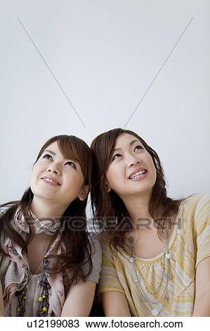 Stock Image - Young women sitting side by side looking up. Fotosearch