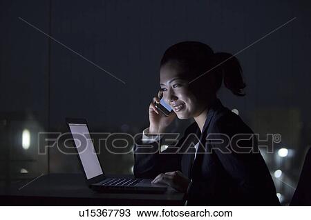 Stock Image - Businesswoman using laptop and smart phone in the dark, Tokyo Prefecture, Honshu, Japan. Fotosearch