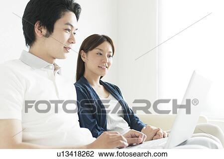 Stock Image - Young couple sitting on sofa using laptop. Fotosearch