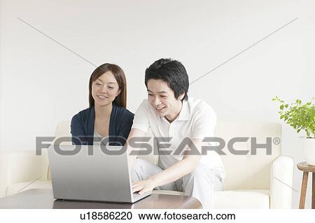 Stock Image - Young couple sitting on sofa using laptop. Fotosearch