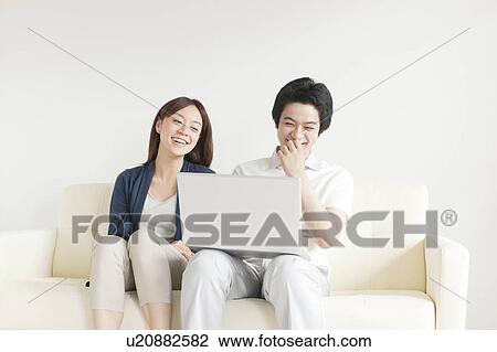 Stock Image - Young couple sitting on sofa using laptop. Fotosearch