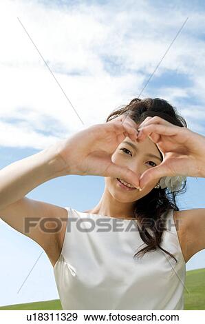 Stock Photo - Young woman wearing a wedding dress making heart shape with hands. Fotosearch