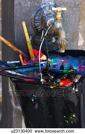 Stock Image - Collection of used paint brushes, spattered with multicoloured, vivid paints, in black plastic bucket hanging on old brass tap. Fotosearch