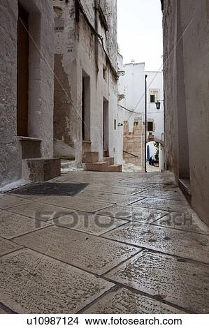 Picture - Street in Ostuni, with typical white stucco walls, stone paving, wrought iron lamp, and archway, with flowers decorating, spring afternoon, Salento, Puglia, Southern Italy. Fotosearch