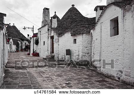 Street view: town of Alberobello, World Heritage site, with trullo houses, white painted stone with piled stone conical roofs, paved street and red flowers set against monochrome view, Puglia, Southern Italy View Large Photo Image Picture - Street view: town of Alberobello, World Heritage site, with trullo houses, white painted stone with piled stone conical roofs, paved street and red flowers set against monochrome view, Puglia, Southern Italy. Fotosearch