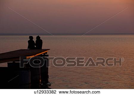 Sunset couple, silhouetted sitting on lakeside dock, Italy View Large Photo Image Stock Image - Sunset couple, silhouetted sitting on lakeside dock, Italy. Fotosearch
