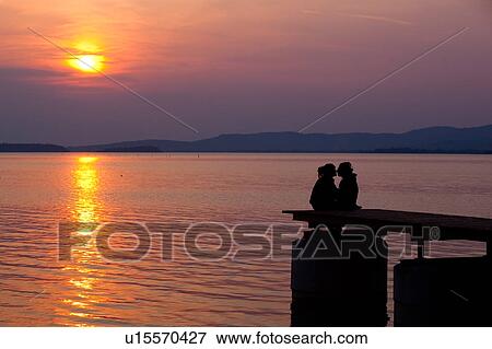 Stock Photo - Sunset couple, silhouetted sitting on lakeside dock, Italy. Fotosearch