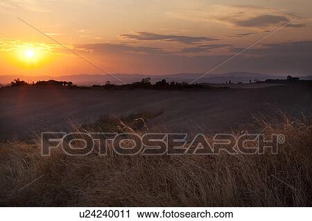 Stock Image - Sunset over rolling hills of Tuscany, with farmland fields in foreground, after harvesting. Fotosearch
