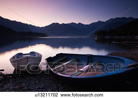 Stock Image - Two traditional wooden fishing boats beached at shore of lake in pink and purple pre-dawn glow, with mountain range and opposite shoreline reflected in calm waters of Lake Como, Northern Italy. Fotosearch