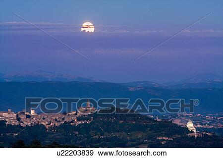 Stock Photo - View of full moon rising over medieval hilltop town of Todi, with spire of San Fortunato, and Tempio of Bramante, town lights illuminated at dusk, Umbria, Italy. Fotosearch