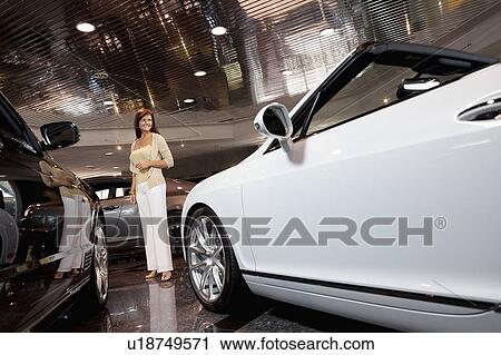 Stock Image - Full-length of smiling woman standing in car showroom. Fotosearch