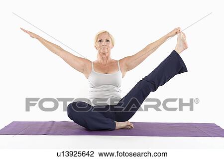 Stock Image - Mature woman sitting on mat and doing yoga with arms outstretched. Fotosearch