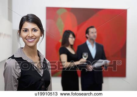 Stock Image - Portrait of smiling young woman in front of a couple in art gallery. Fotosearch