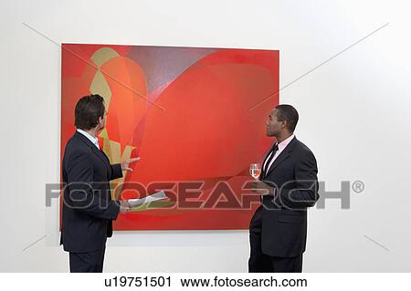Stock Image - Two males talking over painting in art gallery. Fotosearch