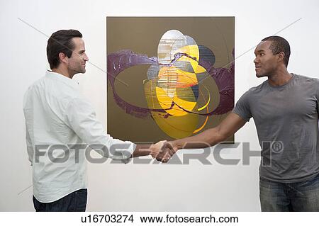 Two man in casuals shaking hands in front of wall painting View Large Photo Image Picture - Two man in casuals shaking hands in front of wall painting. Fotosearch