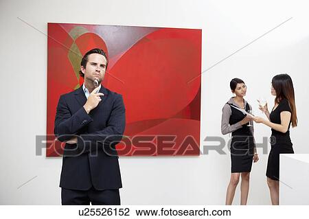 Young man thinking with two women talking in background in art gallery View Large Photo Image Stock Image - Young man thinking with two women talking in background in art gallery. Fotosearch