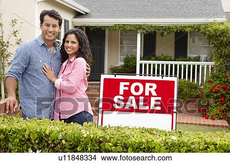 Picture - Hispanic couple outside home with for sale sign. Fotosearch