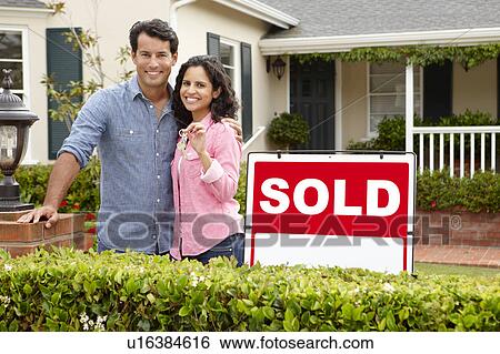 Stock Photograph - Hispanic couple outside home with sold sign. Fotosearch